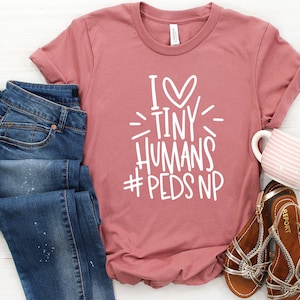 May include: A pink t-shirt with the text "I ♥ Tiny Humans #PedsNP" in white. The shirt is shown with blue jeans and brown sandals.
