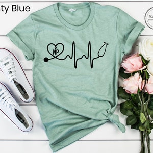 May include: A light green t-shirt with a black graphic of a stethoscope with a heart that says "NP" and a heartbeat line. The t-shirt is on a white surface with white sneakers and pink flowers.