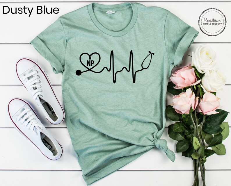 May include: A light green t-shirt with a black graphic of a stethoscope with a heart that says "NP" and a heartbeat line. The t-shirt is on a white surface with white sneakers and pink flowers.