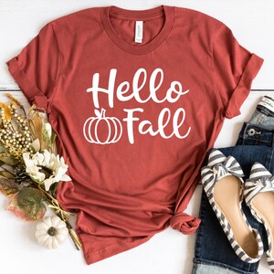 May include: A rust-colored t-shirt with the text "Hello Fall" in white lettering and a white pumpkin graphic. The shirt is laid out on a white wooden surface with a pair of striped flats and blue jeans.