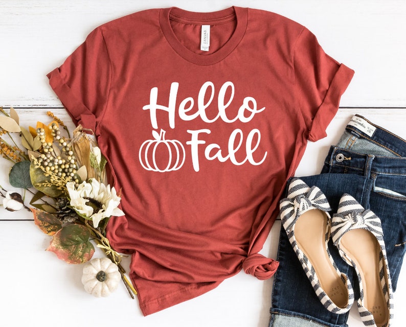 May include: A rust-colored t-shirt with the text "Hello Fall" in white lettering and a white pumpkin graphic. The shirt is laid out on a white wooden surface with a pair of striped flats and blue jeans.