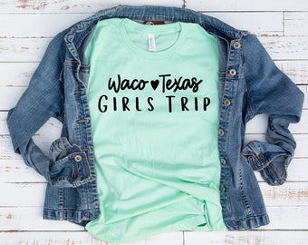 May include: A light green t-shirt with the text "Waco Texas Girls Trip" in black lettering with a heart symbol between "Waco" and "Texas". The t-shirt is folded and laid on a white wooden surface with a blue denim jacket draped over it.