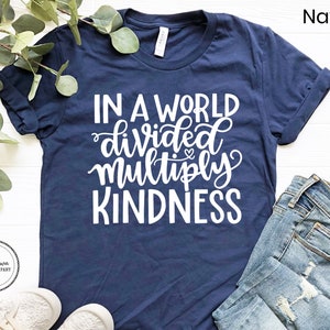 May include: A navy blue t-shirt with the text "In a world divided multiply kindness" in white lettering. The shirt is photographed on a white surface with a sprig of greenery and a pair of white shoes.