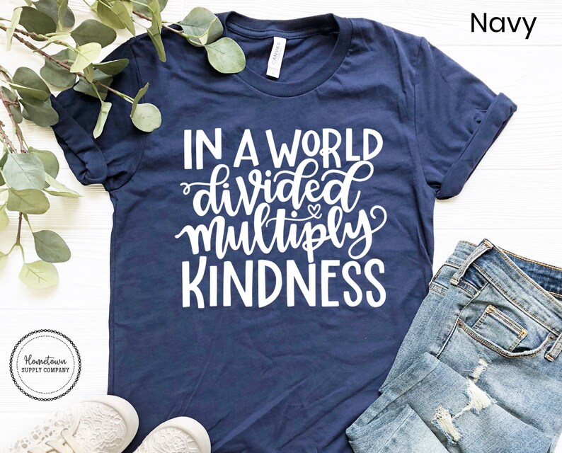 May include: A navy blue t-shirt with the text "In a world divided multiply kindness" in white lettering. The shirt is photographed on a white surface with a sprig of greenery and a pair of white shoes.