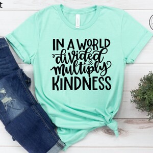 May include: A mint green t-shirt with the text "In a world divided multiply kindness" in black lettering.