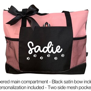 Puede incluir: Bolso de mano rosa y negro con un lazo de sat&eacute;n negro. El bolso tiene un compartimento principal con cremallera, dos bolsillos laterales de malla y un nombre personalizado "Sadie" con huellas de patas.