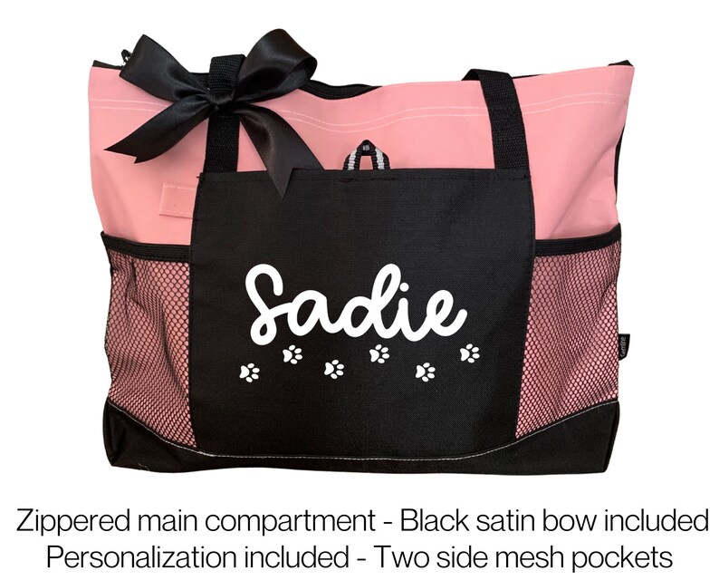Puede incluir: Bolso de mano rosa y negro con un lazo de sat&eacute;n negro. El bolso tiene un compartimento principal con cremallera, dos bolsillos laterales de malla y un nombre personalizado "Sadie" con huellas de patas.