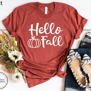 May include: A rust-colored t-shirt with the text "Hello Fall" in white lettering and a white pumpkin graphic. The shirt is laid flat on a white surface with a pair of striped flats and blue jeans.