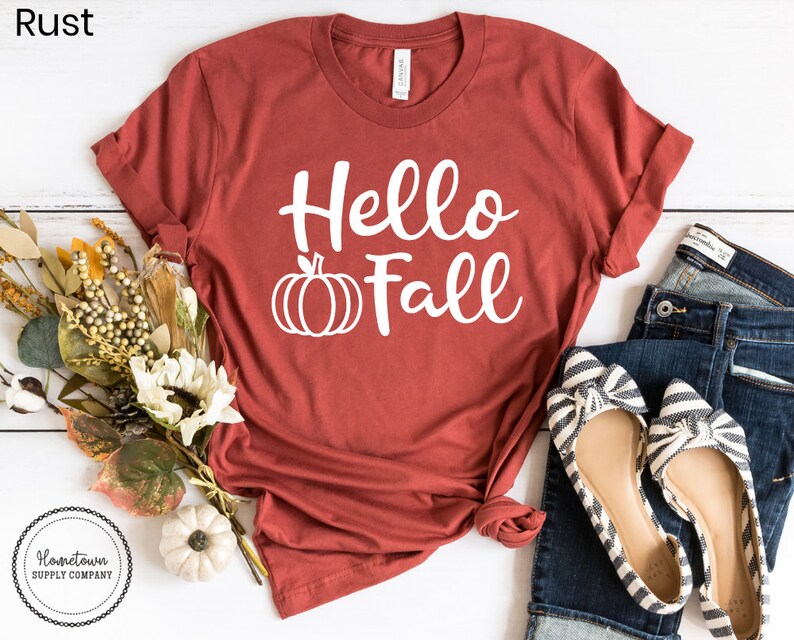 May include: A rust-colored t-shirt with the text "Hello Fall" in white lettering and a white pumpkin graphic. The shirt is laid flat on a white surface with a pair of striped flats and blue jeans.