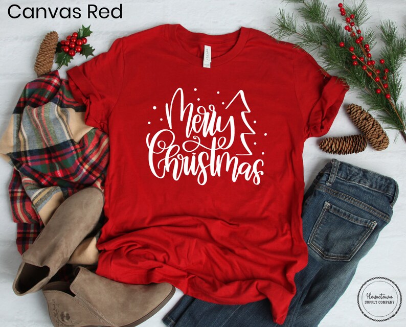Puede incluir: Una camiseta roja con texto blanco que dice "Feliz Navidad" con un gr&aacute;fico de &aacute;rbol de Navidad estilizado.