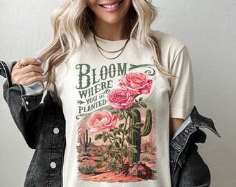 May include: A beige t-shirt with a vintage-style graphic print of pink roses and a cactus. The text on the shirt reads "Bloom Where You Are Planted".