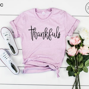 Puede incluir: Camiseta lila con la palabra "thankful" en escritura negra con un dise&ntilde;o de cruz y hoja. La camiseta est&aacute; colocada sobre una superficie de madera blanca con zapatillas blancas y rosas rosas.