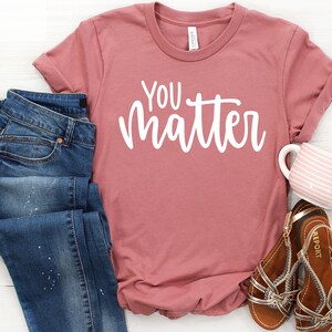 May include: A dusty rose colored t-shirt with the text "YOU matter" in white lettering. The shirt is laid flat on a white wooden surface with a pair of blue jeans, a pair of brown sandals, and a pink and white striped mug.