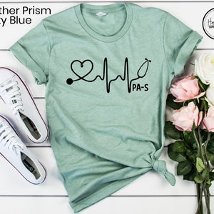 K&ouml;nnte beinhalten: Ein T-Shirt in der Farbe Hellblau mit einem schwarzen Aufdruck eines Stethoskops und eines Herzschlags mit dem Text "PA-S".