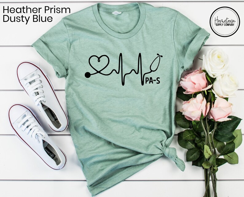 K&ouml;nnte beinhalten: Ein T-Shirt in der Farbe Hellblau mit einem schwarzen Aufdruck eines Stethoskops und eines Herzschlags mit dem Text "PA-S".