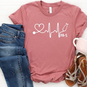 May include: A pink t-shirt with a white graphic of a heart, a stethoscope, and a heartbeat line. The text "PA-S" is below the graphic.