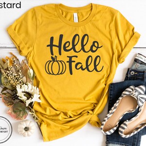 May include: A mustard yellow t-shirt with the text "Hello Fall" in black script and a black pumpkin graphic.