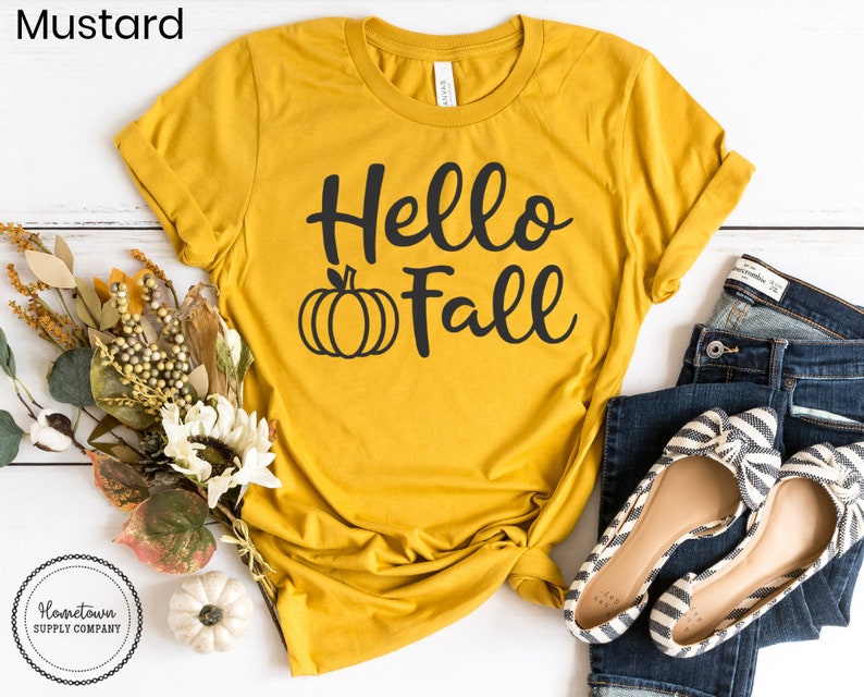May include: A mustard yellow t-shirt with the text "Hello Fall" in black script and a black pumpkin graphic.