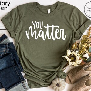 May include: A green t-shirt with the text "YOU matter" in white lettering. The shirt is laid flat on a white surface with a pair of blue jeans, a small pumpkin, and a bouquet of fall flowers.