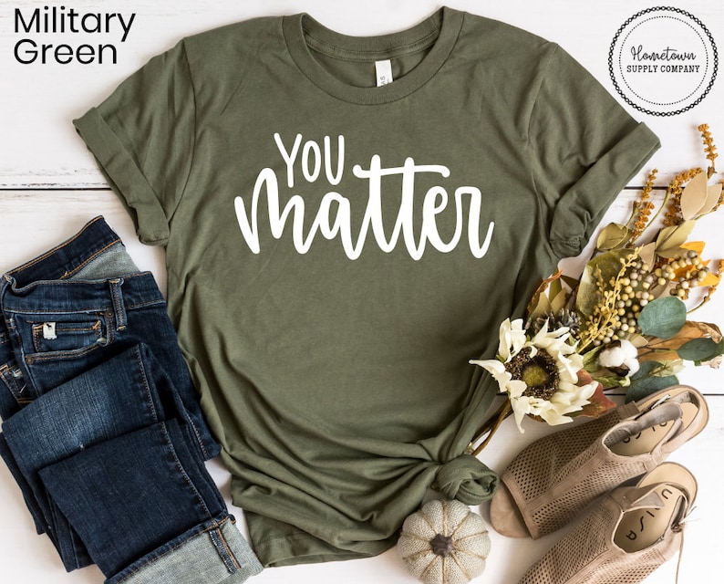 May include: A green t-shirt with the text "YOU matter" in white lettering. The shirt is laid flat on a white surface with a pair of blue jeans, a small pumpkin, and a bouquet of fall flowers.
