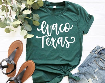 Puede incluir: Una camiseta verde con texto blanco que dice "Waco Texas" en una fuente cursiva. La camiseta está colocada sobre una superficie blanca con un par de sandalias marrones y unos vaqueros azules.