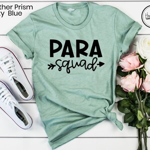 May include: A light green t-shirt with the text "PARA squad" in black lettering. The shirt is laid flat on a white surface with a bouquet of white and pink roses to the right.