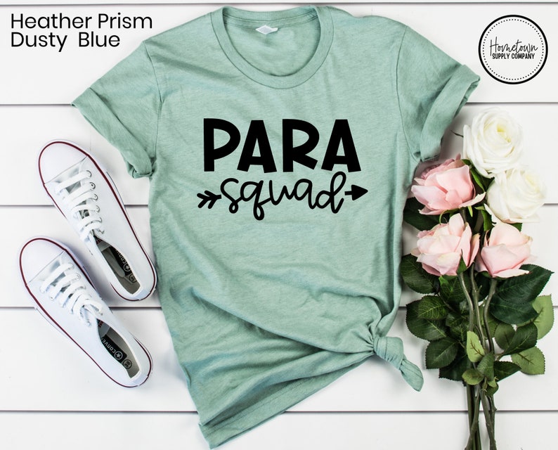 May include: A light green t-shirt with the text "PARA squad" in black lettering. The shirt is laid flat on a white surface with a bouquet of white and pink roses to the right.
