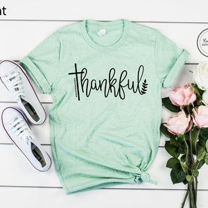 Puede incluir: Una camiseta verde menta con la palabra "thankful" impresa en negro. La camiseta est&aacute; doblada y colocada sobre una superficie de madera blanca. Hay zapatillas blancas y flores rosas en el fondo.