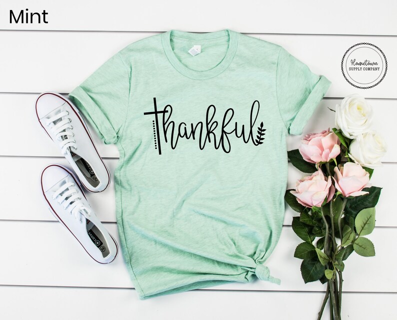 Puede incluir: Una camiseta verde menta con la palabra "thankful" impresa en negro. La camiseta est&aacute; doblada y colocada sobre una superficie de madera blanca. Hay zapatillas blancas y flores rosas en el fondo.