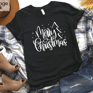 Puede incluir: Camiseta negra con un gr&aacute;fico blanco que dice "Feliz Navidad" con un &aacute;rbol de Navidad estilizado.