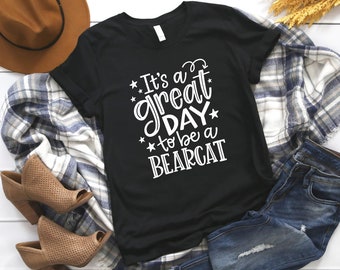 Puede incluir: Camiseta negra con texto blanco que dice "It's a great day to be a Bearcat". La camiseta está colocada sobre una superficie blanca con una manta a cuadros, un sombrero marrón y unos vaqueros azules.
