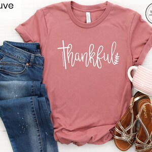 Puede incluir: Camiseta de color malva con la palabra "thankful" en letras blancas. La camiseta se combina con unos vaqueros azules y unas sandalias marrones.