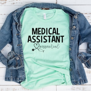 May include: A light green t-shirt with the text "MEDICAL ASSISTANT #essential" in black lettering. A stethoscope with a heart is above the text. The t-shirt is folded and laid on a white wooden surface.