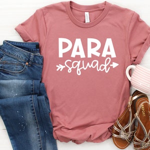 May include: A dusty rose t-shirt with the text "PARA squad" in white. The shirt is laid flat on a white wooden surface with a pair of blue jeans, a mug, and sandals.