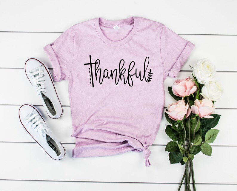 Puede incluir: Una camiseta rosa claro con la palabra "thankful" escrita en cursiva negra. La palabra est&aacute; decorada con una peque&ntilde;a cruz y una hoja a cada lado. La camiseta est&aacute; colocada sobre una superficie de madera blanca con un ramo de rosas rosas y dos zapatillas blancas.