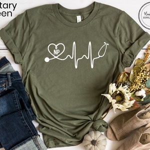 May include: A military green t-shirt with a white graphic of a stethoscope, a heart with the letters "NP" inside, and a heartbeat line. The t-shirt is on a white surface with a pair of brown sandals and a small pumpkin.