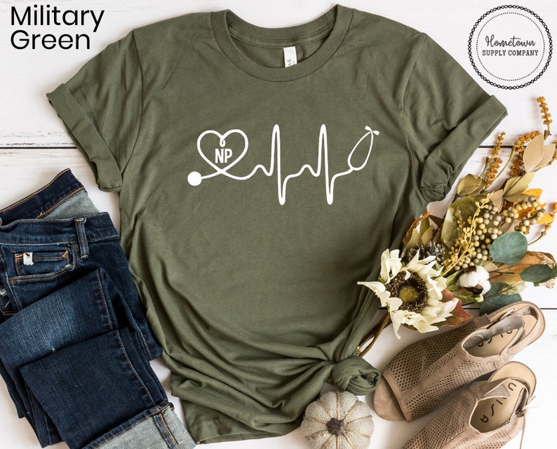 May include: A military green t-shirt with a white graphic of a stethoscope, a heart with the letters "NP" inside, and a heartbeat line. The t-shirt is on a white surface with a pair of brown sandals and a small pumpkin.