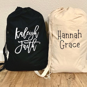 May include: Two drawstring laundry bags, one black with white text "Kaleigh Faith" and one beige with black text "Hannah Grace".