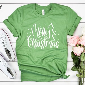 Puede incluir: Camiseta verde con texto blanco que dice "Feliz Navidad" con un gr&aacute;fico de &aacute;rbol de Navidad estilizado.