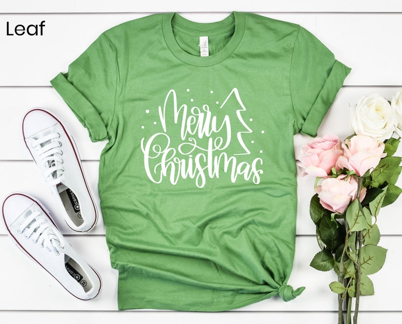 Puede incluir: Camiseta verde con texto blanco que dice "Feliz Navidad" con un gr&aacute;fico de &aacute;rbol de Navidad estilizado.