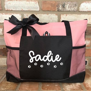 Puede incluir: Bolsa de mano rosa y negra con un lazo negro y el nombre "Sadie" en blanco con huellas de patas en la parte delantera.