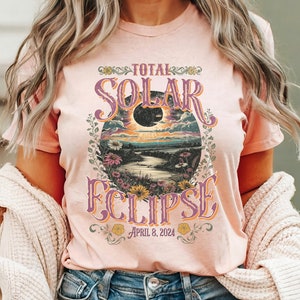 May include: A pink t-shirt with a vintage-style graphic of a total solar eclipse with the text "TOTAL SOLAR ECLIPSE APRIL 8, 2024".