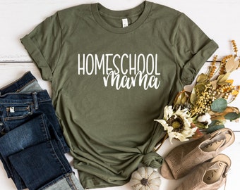 May include: A green t-shirt with the text "HOMESCHOOL mama" in white lettering. The shirt is folded and laid on a white wooden surface with a pair of jeans, a small white pumpkin, and a bouquet of dried flowers.