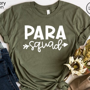 May include: A green t-shirt with the text "PARA squad" in white lettering. The shirt is on a white wooden surface with a pair of jeans and a bouquet of flowers.