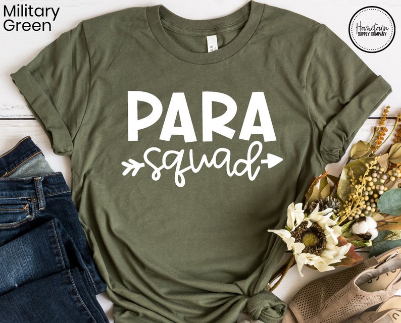 May include: A green t-shirt with the text "PARA squad" in white lettering. The shirt is on a white wooden surface with a pair of jeans and a bouquet of flowers.