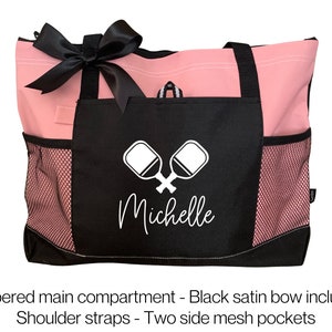 May include: Pink and black tote bag with a zippered main compartment, two side mesh pockets, and a black satin bow. The bag has a black pocket with a white paddleball graphic and the name "Michelle" in white text.