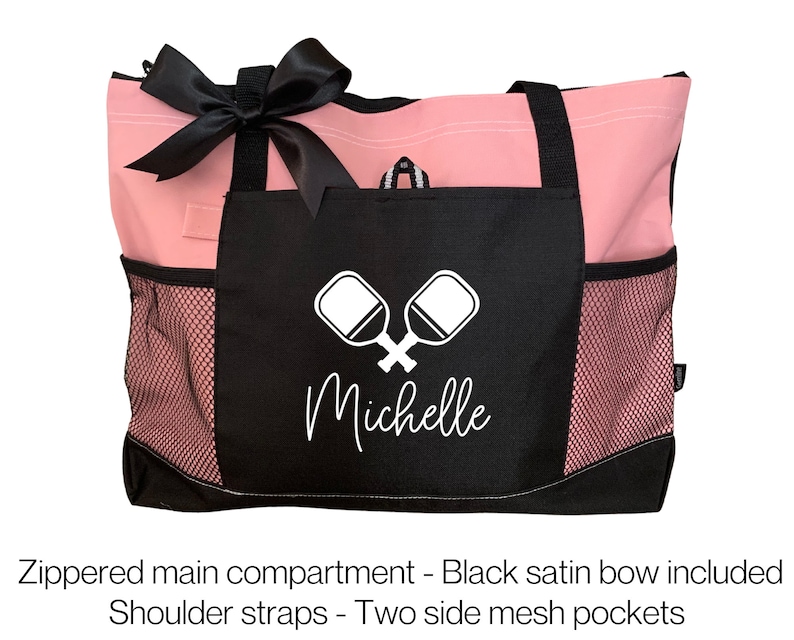 May include: Pink and black tote bag with a zippered main compartment, two side mesh pockets, and a black satin bow. The bag has a black pocket with a white paddleball graphic and the name "Michelle" in white text.