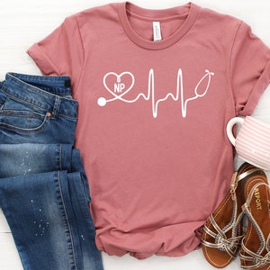 May include: A pink t-shirt with a white graphic of a stethoscope with a heart that says "NP" and a heartbeat line.