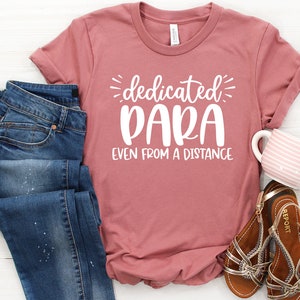 May include: A pink t-shirt with the text "dedicated para even from a distance" in white lettering. The shirt is shown with blue jeans and brown sandals.