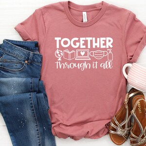 May include: A pink t-shirt with the text "TOGETHER through it all" in white. The text is accompanied by a globe, a book, a laptop with a heart, a face mask, and a bottle of hand sanitizer.